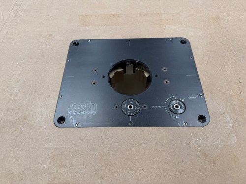 Close-up view of a JessEm router lift mounted on a flat surface, featuring circular opening and adjustment knobs.