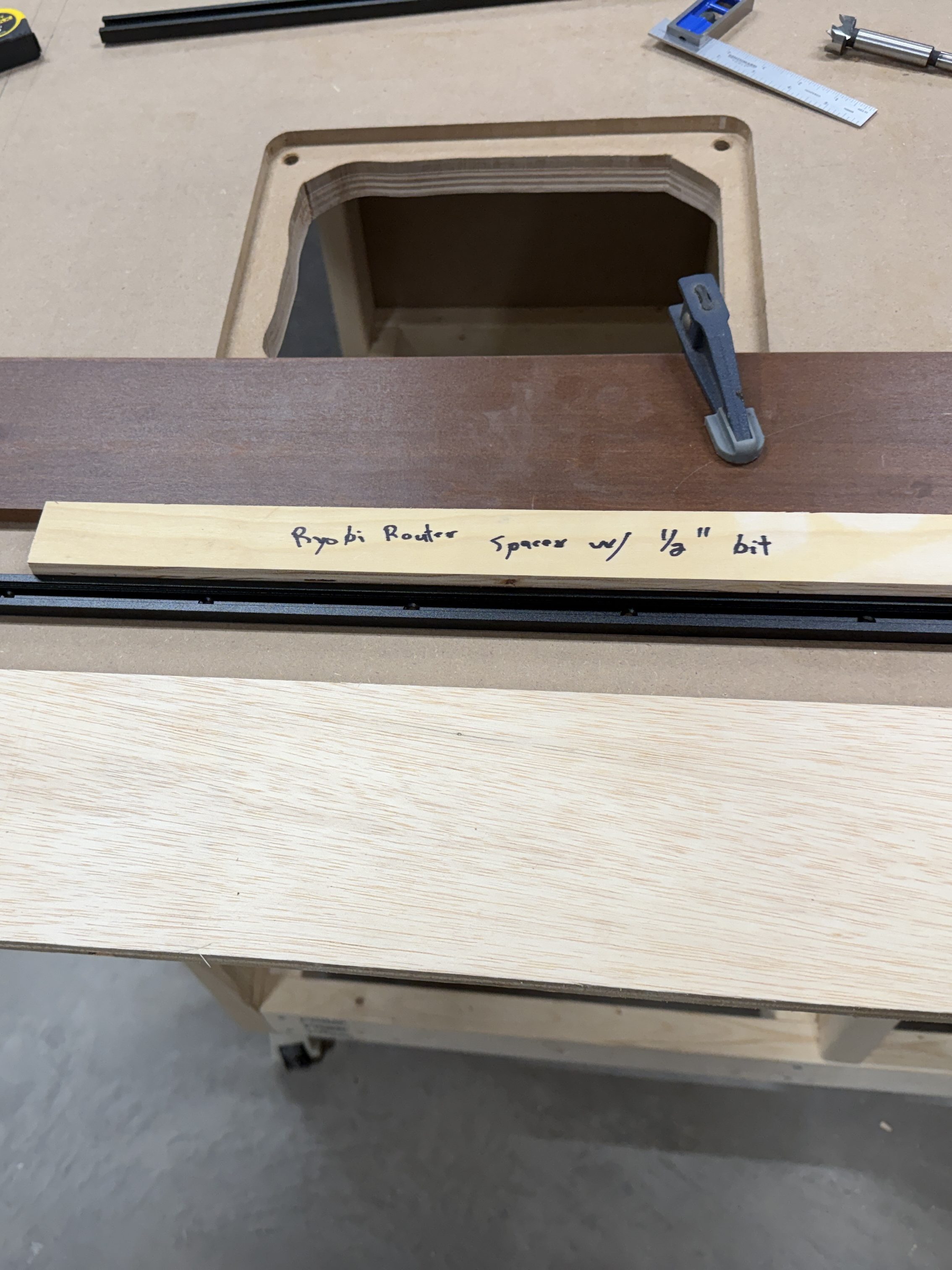 Close-up of a router station setup showing a spacer labeled 'Ryobi Router Spacer w/ 1/8" bit' positioned on a wooden work table with T-tracks in place.