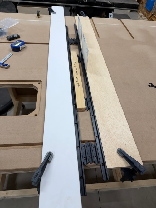 Close-up of a router table setup showing a T-track and fence alignment, with clamps securing components on a work surface.