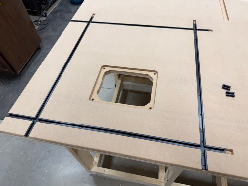 A close-up of a router table setup showing T-tracks being installed around a cutout for the router lift on a work surface.