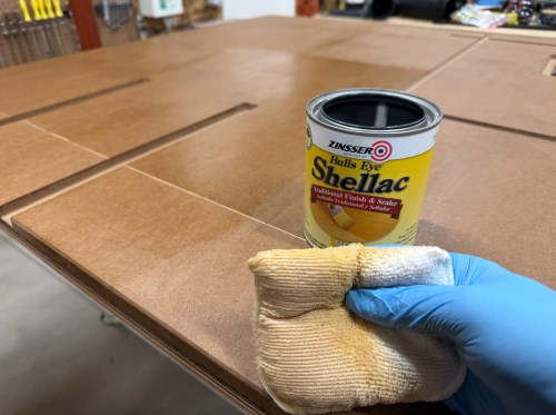 A person applying Zinsser Bulls Eye Shellac on a wood surface with a cloth, wearing a blue glove.