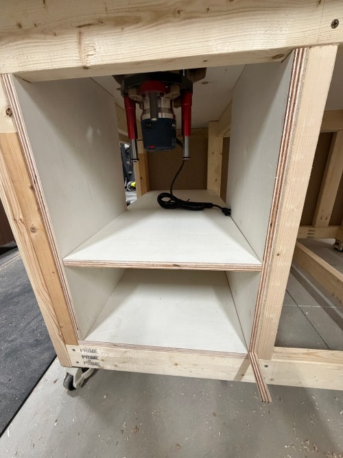 View of a router station cabinet with a Bosch router mounted above a shelf, showing the interior structure and power cord.