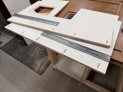 A workbench with plywood pieces laid out, showing a cut-out section and drawer slides positioned along the top.