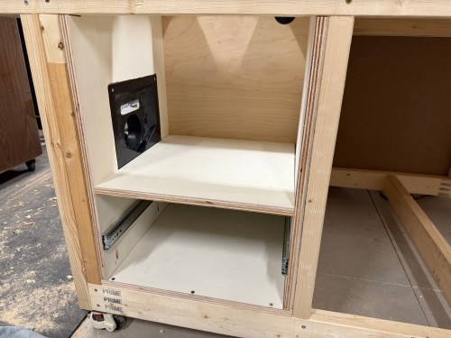Interior view of a wooden cabinet for a router station, featuring a mounted dust collection port and a shelf.