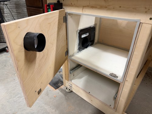 A wooden cabinet with a door open, showing a dust collection system inside and an attached hose on the side.