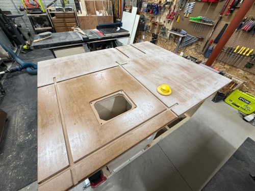 A wooden table surface featuring a routed hole for a router lift, with T-track outlines drawn on it and a yellow measuring tool positioned nearby.