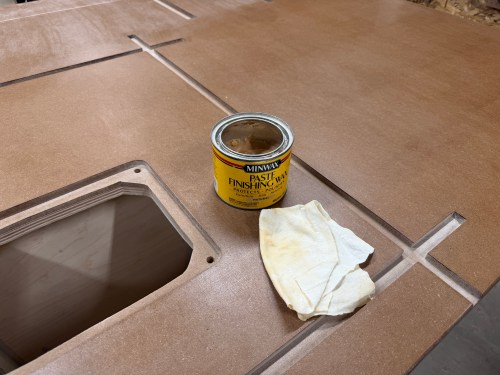 A can of paste finishing wax and a cleaning cloth placed on a router table surface, featuring a cutout for the router lift.