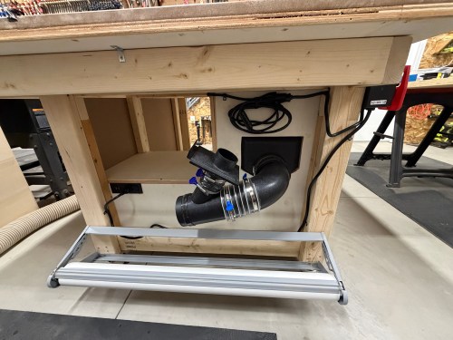 View of the underside of a woodworking table showing the dust collection system with a hose and various connectors.
