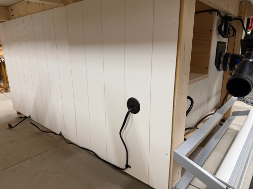 View of the side of a router table cabinet showing a white panel with a power cord and dust collection hose exiting the side.