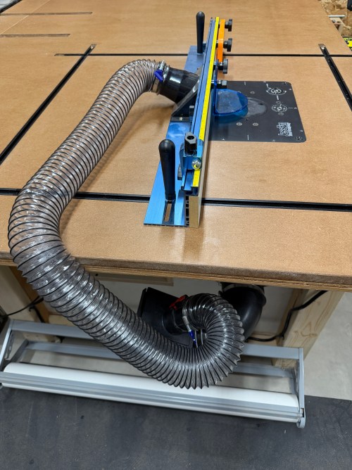A router station equipped with a dust collection system, including a clear hose connected to a dust hood, mounted on a wooden table with a fence and control panel.