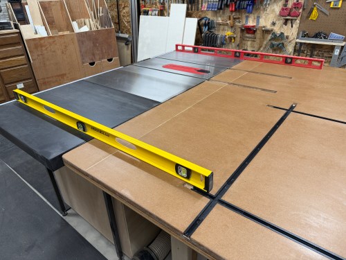 A woodworking table with a level placed on top, showing the setup for a router station, including a saw and T-track system in a workshop.