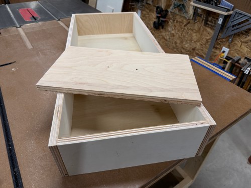 Close-up of a partially assembled wooden drawer with a lid resting on top, placed on a workbench in a workshop.