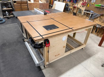 A woodworking router table setup featuring a large wooden surface with T-tracks, a mounted router lift, and a dust collection system. The table includes various tools and materials organized on the surrounding wall.