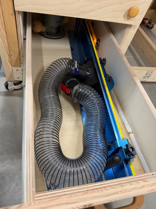 A close-up view of a drawer in a router station, featuring a clear dust collection hose positioned next to a blue T-track with measurement markings.